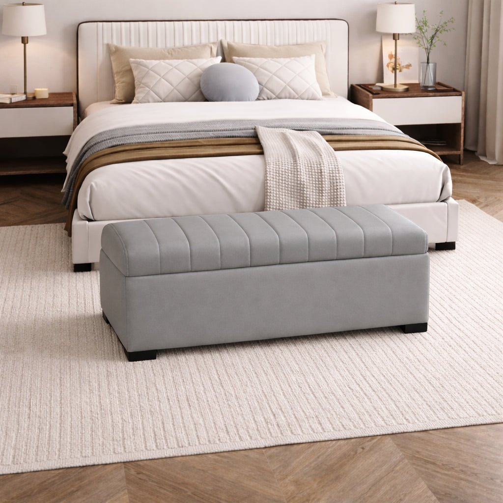 Gray tufted ottoman in front of a neatly made bed in a bedroom setting.