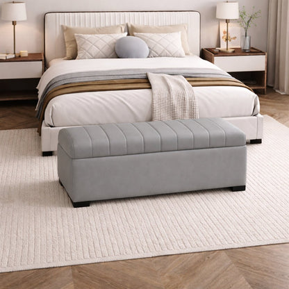 Gray tufted ottoman in front of a neatly made bed in a bedroom setting.