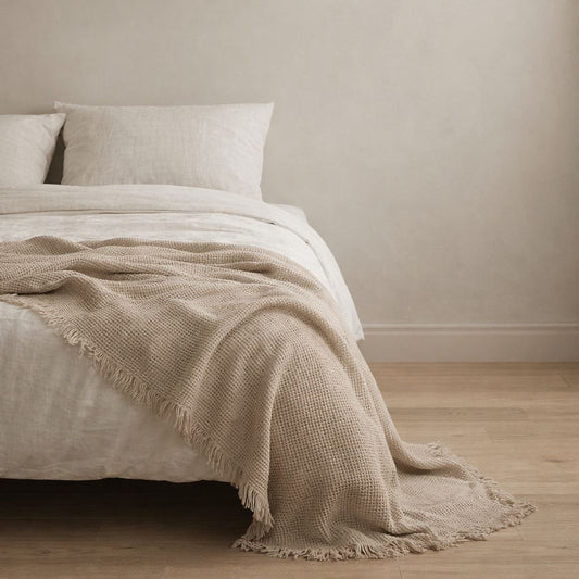 Beige blanket draped over a bed with white pillows against a beige wall.